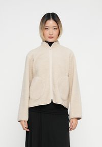 Νέος άνθρωπος ντυμένος με μπεζ φούτερ με φερμουάρ και μεγάλα μπροστινά τσέπες, φορώντας μαύρα ρούχα από κάτω, που στέκεται σε λευκό φόντο.