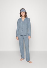Blå pyjamasset med prickar, tillverkat av mjukt material, med lång ärm, knäppning och matchande byxor. Inkluderar en koordinerande ögonmask.