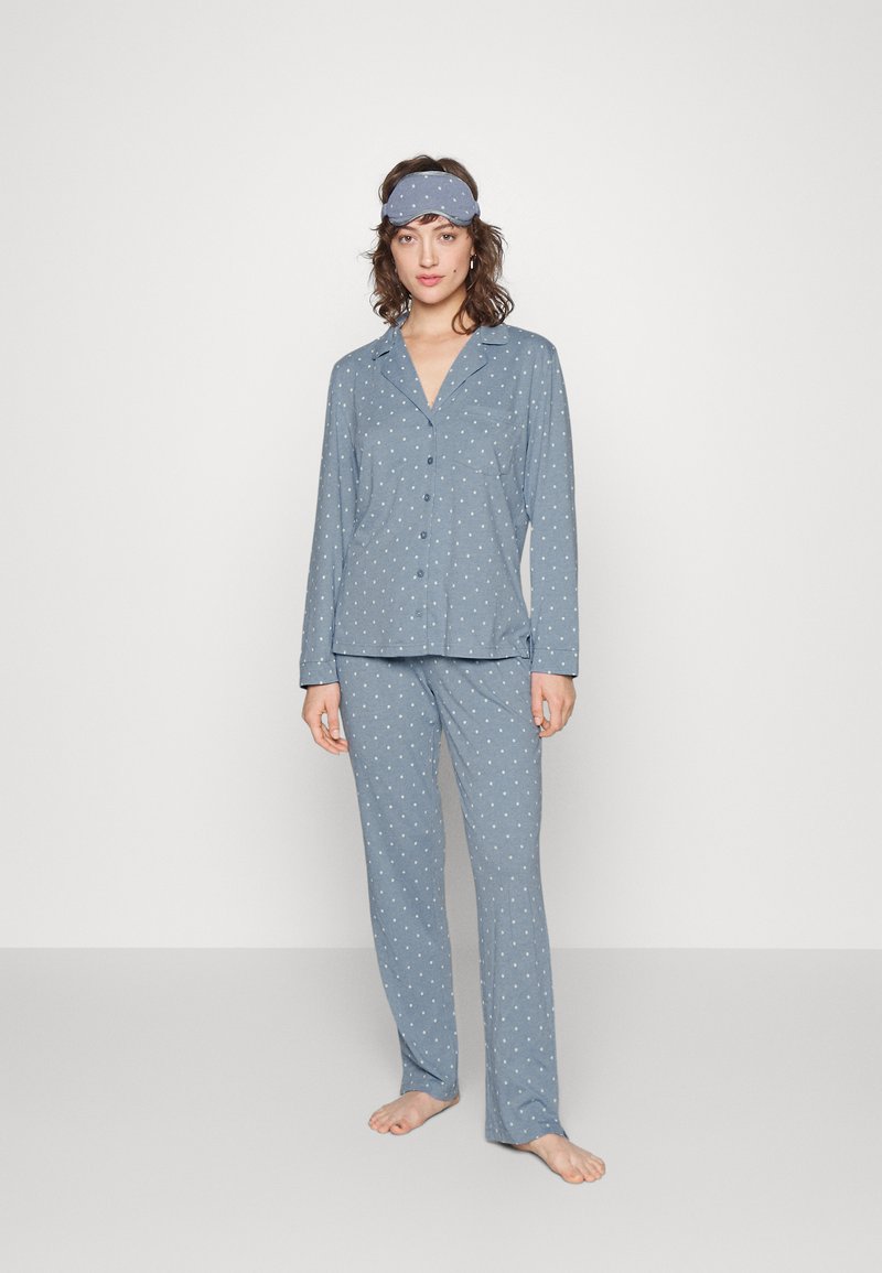Blå pyjamasset med prickar, tillverkat av mjukt material, med lång ärm, knäppning och matchande byxor. Inkluderar en koordinerande ögonmask.