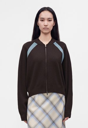 Femme aux longs cheveux noirs porte un pull zippé marron avec des touches bleues aux épaules et une jupe à carreaux beige et bleue sur un fond uni.