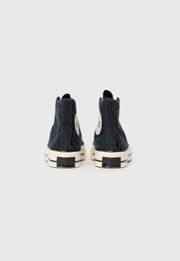 Adidași înalți, textura neagră, cu talpă de cauciuc albă și patch cu logo. Au o parte superioară cu un model și accente contrastante în culoare crem.