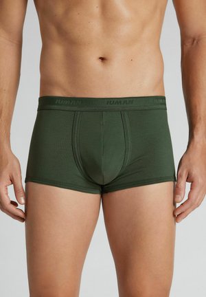 Männlicher Oberkörper, der dunkelgrüne, enganliegende Boxershorts mit einer genähten Fronttasche und elastischem Bund vor neutralem Hintergrund trägt.