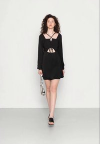 Mulher com cabelo cacheado, vestindo um vestido preto com recortes, segurando uma bolsa branca, caminhando para a frente contra um fundo branco liso.