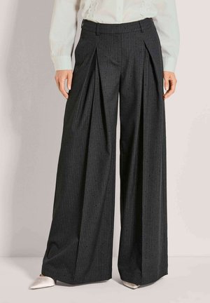 Schwarze, weit geschnittene Nadelstreifenhose mit vorderen Falten, getragen zu weißer Bluse und weißen spitzen Schuhen.