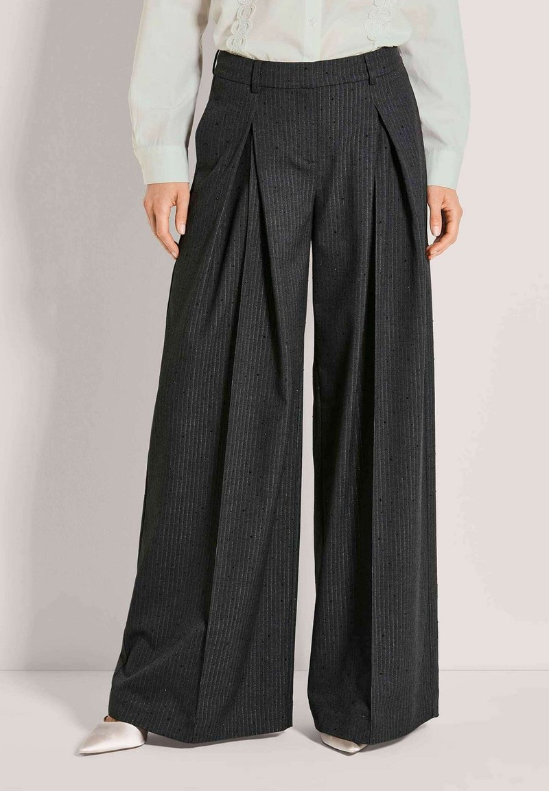 Schwarze, weit geschnittene Nadelstreifenhose mit vorderen Falten, getragen zu weißer Bluse und weißen spitzen Schuhen.