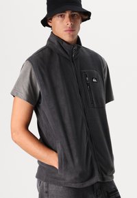 Gray fleece vest with a high collar, zip front, and a chest pocket. Worn over a gray short-sleeve shirt, featuring side pockets.