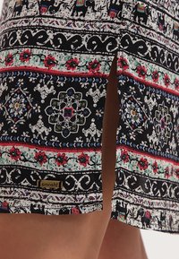 Tissu noir à motifs avec des éléphants et des fleurs répétés, avec une fente sur le côté pour le mouvement, étiquette de marque à l'ourlet.
