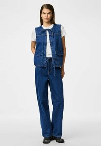 Traper prsluk s vezivanjem, s dva prednja džepa, uparen s širokim traper hlačama. Oba komada su u srednjem plavom pranju.