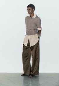 Taupe close-fitting top met opgerolde mouwen, gelaagd over een lichtbeige shirt; brede bruine satijnen broek; zwarte sandalen; minimalistisch ontwerp.