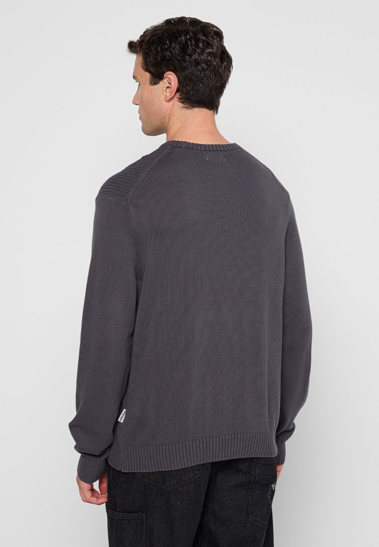 Man in een donkergrijze gebreide trui met lange mouwen en zwarte broek, met de rug naar de camera gericht tegen een effen lichtgrijze achtergrond.