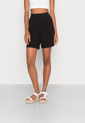 Femme portant un short noir taille haute et ajusté, ainsi que des sandales blanches à plateforme, debout avec les bras détendus le long du corps.