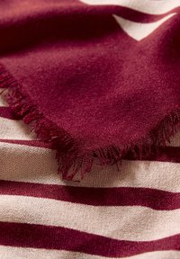 Close-up af et foldet bordeaux halstørklæde med frynser, som ligger på en beige og bordeaux stribet stofoverflade.