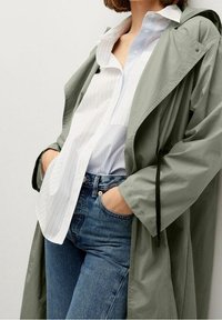 Lichtgewicht olijfgroene regenjas met een losse pasvorm, voorzien van een capuchon met trekkoord, gecombineerd met een wit gestreept overhemd met knopen en blauwe spijkerbroek.