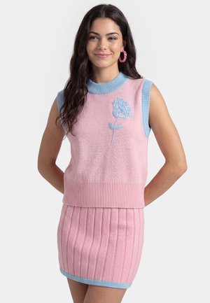 Ung kvinde med langt mørkt hår bærer en lyserød ærmeløs sweater med blåt blomsterdesign og matchende lyserød ribstrikket nederdel med blå kant.