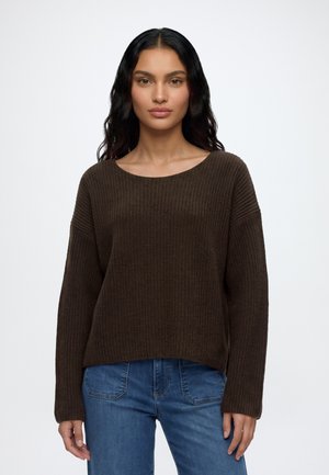Femme aux longs cheveux foncés portant un pull en maille manches longues marron foncé et un jean bleu, debout sur un fond blanc uni.