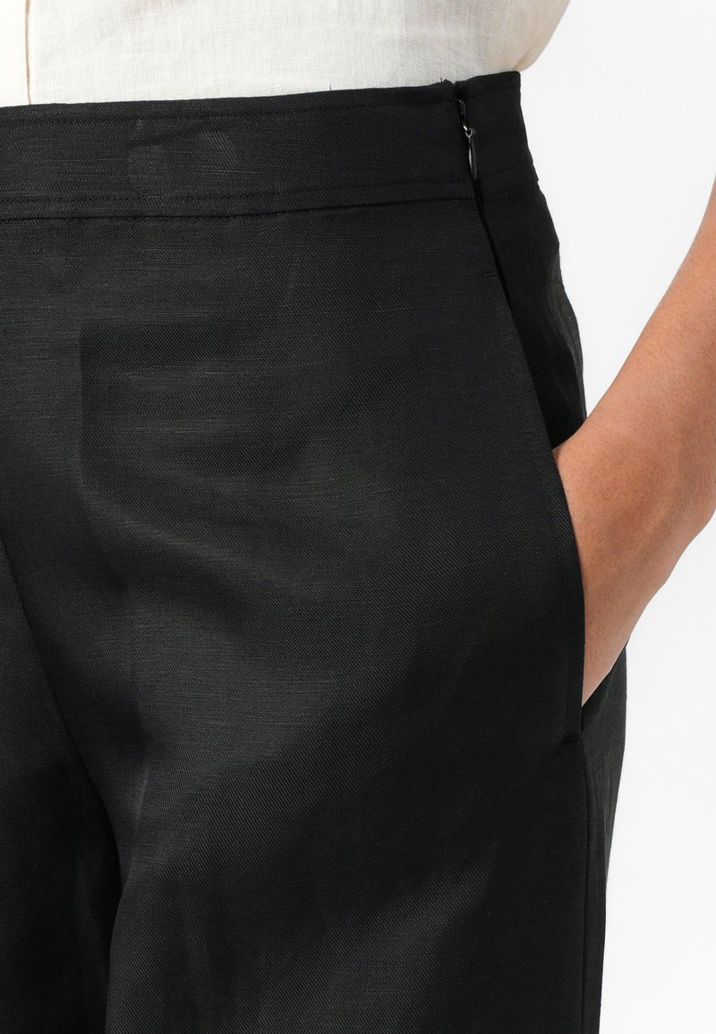 Schwarze Hose mit glatter Textur, ausgestattet mit einem Reißverschluss und Seitentaschen. Der Bund ist maßgeschneidert und sitzt knapp über der Hüfte.