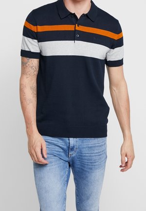 Homme portant un polo bleu marine avec des rayures horizontales orange et grises, associé à un jean bleu clair, debout les bras détendus.