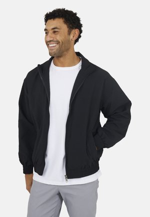 Uśmiechnięty mężczyzna w czarnej kurtce na zamek, białej koszulce i jasnoszarych spodniach, stojący z jedną ręką w kieszeni.