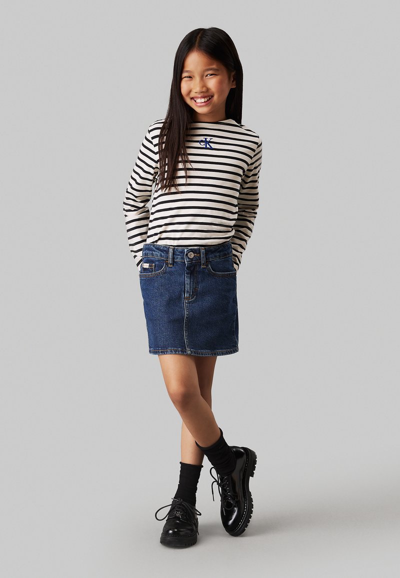 Camicia a maniche lunghe a righe beige e nera, abbinata a una gonna di jeans. Scarpe nere stringate e calzini neri completano l'outfit.