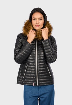 Vrouw in zwarte gewatteerde jas met een bruine met bont gevoerde capuchon, die de capuchon bij haar gezicht houdt, gekleed in blauwe broek tegen een effen achtergrond.