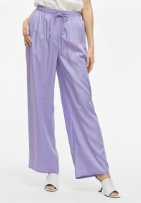 Lichtpaarse wijde broek met een elastische tailleband en trekkoorddetail, gemaakt van een gladde, glanzende stof, gecombineerd met witte open teen hakken.