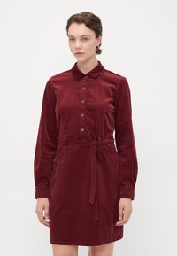 Burgundijas krāsas kordurowa kleita ar apkakli, pogu priekšpusi, garām piedurknēm un sasietu jostu. Iespējamas sānu kabatas un mīksta, teksturēta apdare.