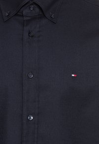 Camicia button-up di un blu navy scuro realizzata in tessuto morbido. Presenta un colletto, una patta frontale con bottoni e un piccolo logo in rosso, bianco e blu.