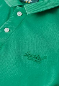 Camisa polo verde hecha de tejido texturizado con cuello tipo polo. Cuenta con dos botones blancos y un logo de marca bordado en el pecho.