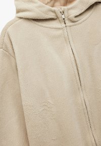 Beige fleece hoodie med en blød tekstur, lynlås lukning og hætte. Har en afslappet pasform og synlige sømdetaljer.