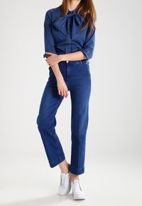 Denimskjorta med en stor fluga-krage, långärmad och med knappstängning, parat med raka blå jeans och vita sneakers.