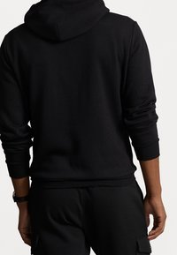 Sudadera negra con capucha, mangas largas, puños acanalados y un ajuste relajado. El material parece suave, con una textura lisa y detalles mínimos.