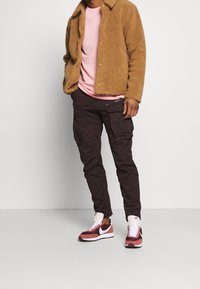 Brun, fluffig-texturerad jacka; ljusrosa T-shirt under; mörka cargo-byxor med dragkedjefickor; röda och vita sneakers.