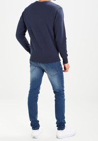 Sweatshirt bleu marine à col rond avec des poignets et une taille côtelés, associé à un jean slim bleu et des baskets blanches. Design simple et de couleur unie.