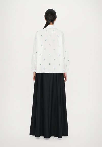 Blouse blanche avec broderie florale verte, associée à une jupe fluide noire. Manches longues avec des poignets élastiques, texture de tissu lisse.