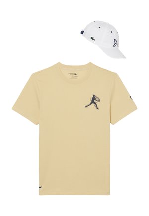 Camiseta beige de Lacoste con un gráfico de silueta en azul marino y gorra blanca de Lacoste con el logo y el número 93 en el lateral.