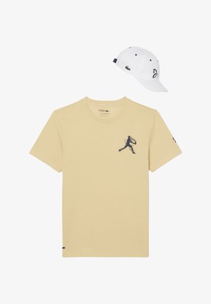 Camiseta beige de Lacoste con un gráfico de silueta en azul marino y gorra blanca de Lacoste con el logo y el número 93 en el lateral.