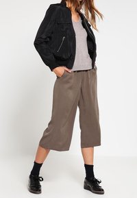 Schwarze Bomberjacke mit Reißverschlusstaschen, graues V-Ausschnitt-Oberteil und weite, graue, verkürzte Hose. Gestylt mit schwarzen Schuhen und dunklen Socken.