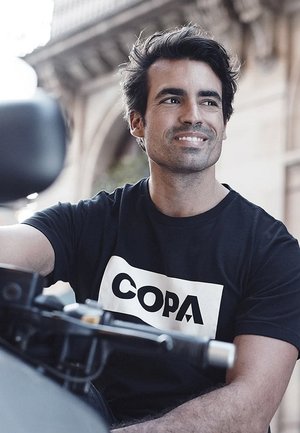Zwarte T-shirt met het "COPA" logo in het wit op de voorkant, zittend op een motorfiets; de achtergrond bevat architectonische elementen.