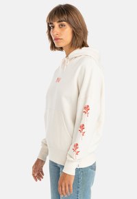 Cream hoodie with red rose embroidery on sleeves, drawstring hood, kangaroo pocket, and ribbed cuffs. Worn with light blue jeans.