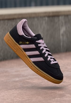 Czarny sneaker Adidas Spezial z różowymi paskami, różowymi sznurowadłami i brązową teksturowaną podeszwą unoszący się nad betonową powierzchnią.