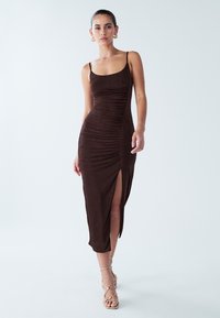 Robe marron à bretelles spaghetti avec une texture froncée et une fente latérale, réalisée dans un tissu doux. Associée à des sandales dorées à brides.