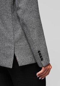 Šedý vzorovaný blazer herringbone s upraveným střihem, čtyřmi černými knoflíkovými detaily na manžetách a rozparkem pro daleko větší volnost pohybu.