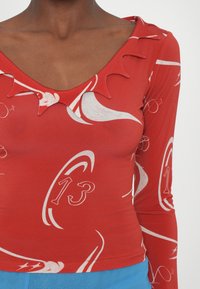 Rød langermet topp med V-hals og intrikate bølgede kanter. Den har et hvitt abstrakt mønster og nummeret "13."