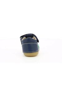 Zapato de piel azul marino con una correa de velcro, detalles de costuras blancas y una suela marrón. Textura suave con un diseño de tacón redondo.