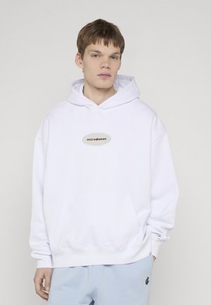 Νέος άνδρας με λευκή μπλούζα Rocawear και ανοιχτό μπλε παντελόνι, στέκεται με το ένα χέρι εν μέρει στην τσέπη του μπροστά σε απλό φόντο.