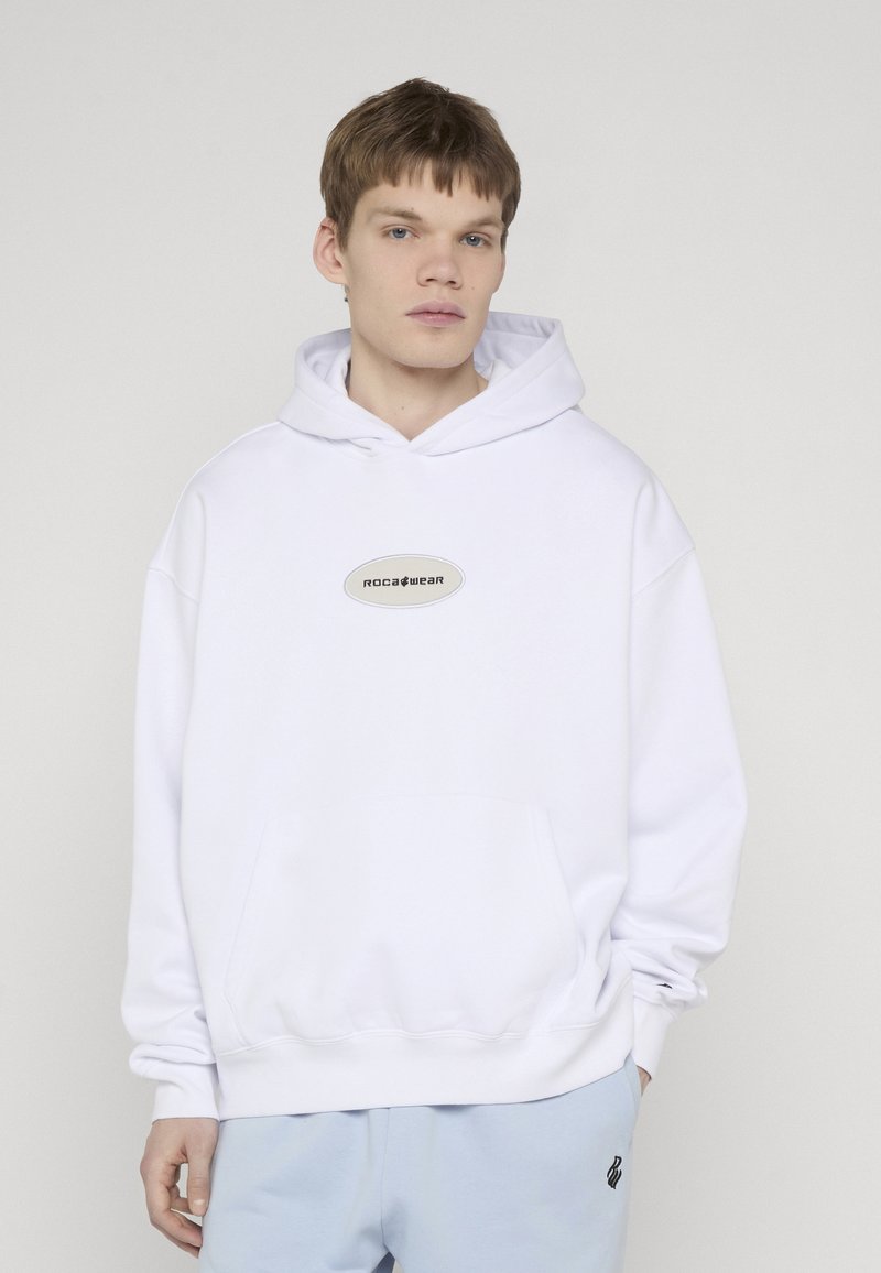 Νέος άνδρας με λευκή μπλούζα Rocawear και ανοιχτό μπλε παντελόνι, στέκεται με το ένα χέρι εν μέρει στην τσέπη του μπροστά σε απλό φόντο.