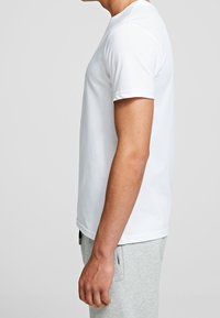 Camiseta blanca de algodón con mangas cortas, cuello redondo y corte regular. Sin patrones ni gráficos visibles, combinada con pantalones de chándal grises.
