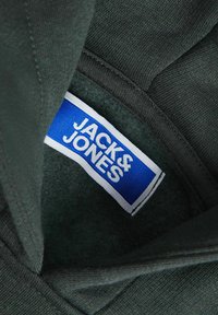 Tmavozelená tkanina vystavuje značku JACHS & JONES v modrej a bielej farbe vo vnútri výstrihu odevu, s textúrovaným povrchom.