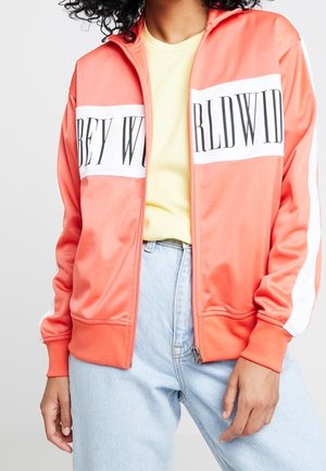 Osoba ubrana w koralową kurtkę na zamek z białym paskiem i czarnym napisem "OBEY WORLDWIDE", w połączeniu z żółtą koszulką i jasnoniebieskimi dżinsami.