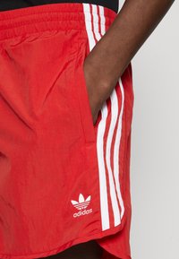 Shorts Adidas roșii, cu o textură netedă, având dungi laterale albe și logo, concepuți cu un buzunar pe partea dreaptă.